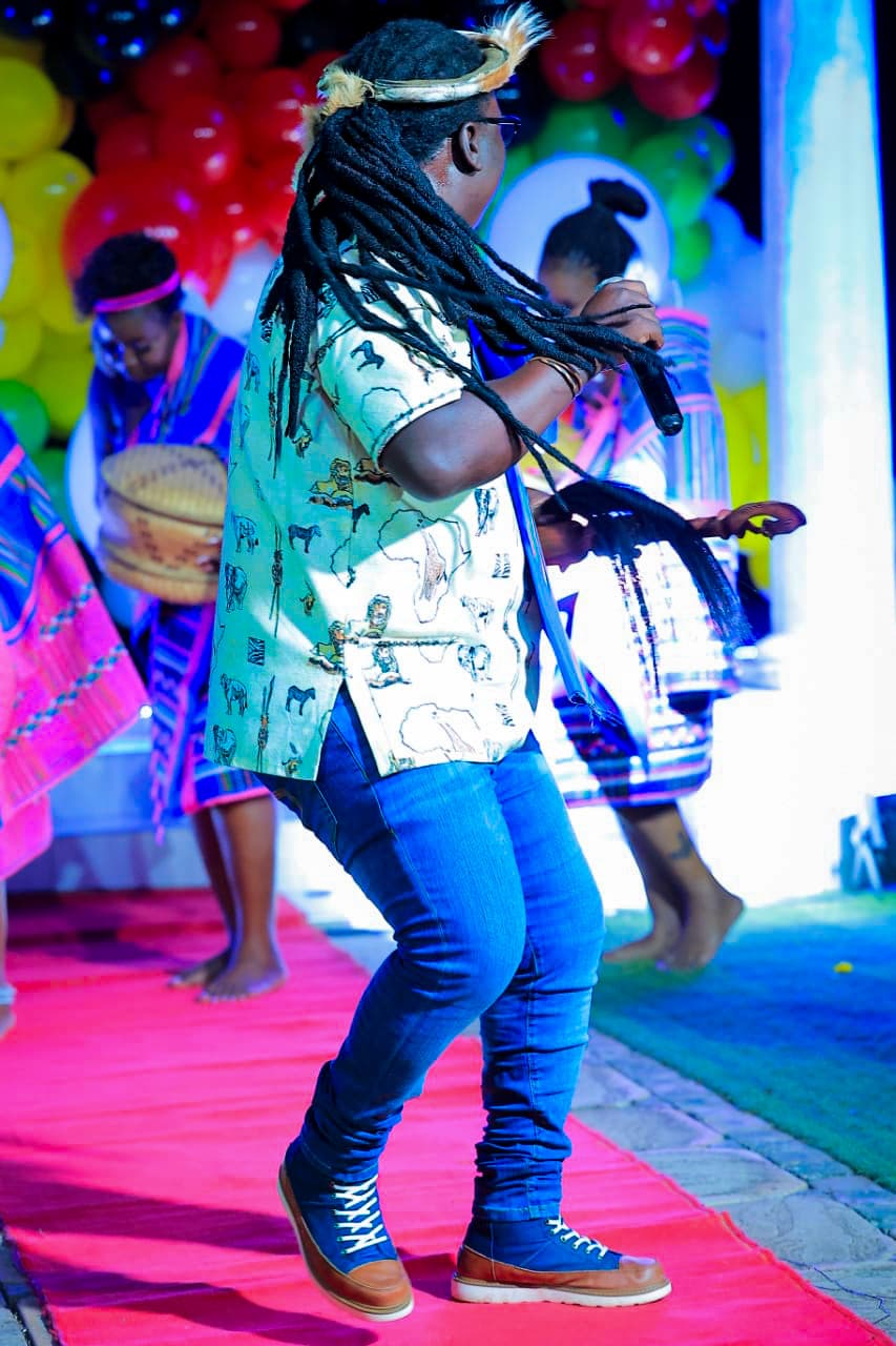 Papa Thile mid-movement on a lit stage, long locs swinging, seen from behind and to the side while holding a microphone. Dancers in Venda dress and a balloon backdrop surround the pink stage carpet.